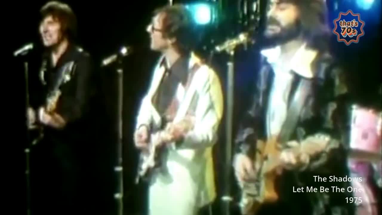 Three men stand on a stage, each holding a guitar. The lead singer, in a black shirt, belts into a microphone. Behind them, bright stage lights illuminate the scene.