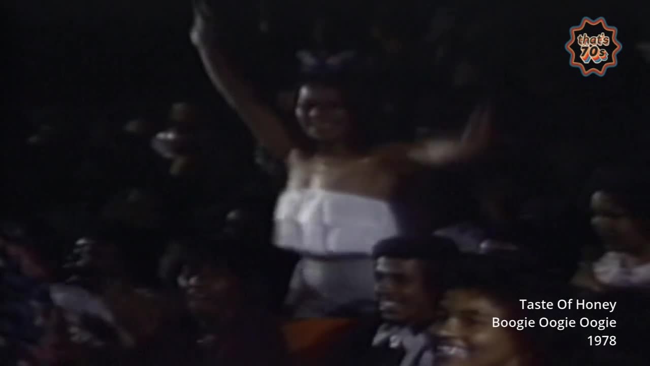 A woman in a white ruffled top throws her arms up in the air, her face beaming. The crowd around her is a sea of dark shapes, all caught up in the energy of the moment.