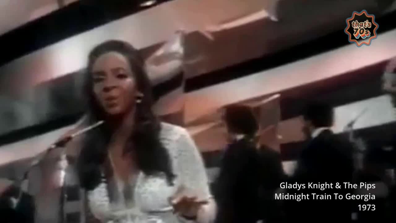 Gladys Knight, in a sparkly white dress, belts out a tune. Behind her, her bandmates in dark suits are visible.