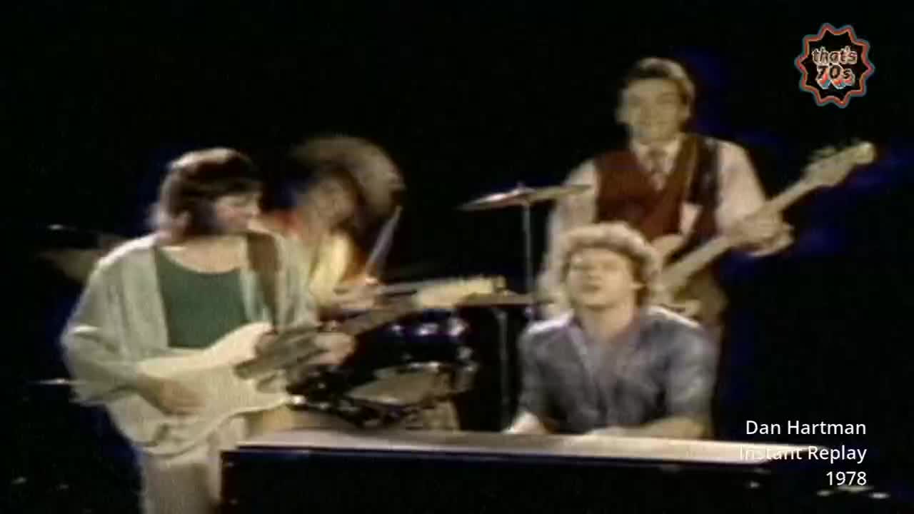 A band plays on stage, the guitarist on the left wearing a light cardigan over a green shirt. The singer in the foreground looks up, his curly hair catching the light.