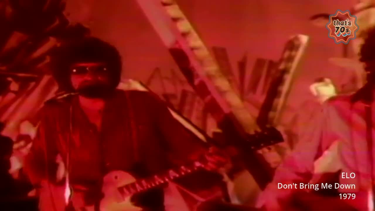 A man with an afro and sunglasses plays a white electric guitar, his fingers moving across the fretboard. The stage is bathed in a deep red light, with angular, abstract shapes looming behind the musicians.