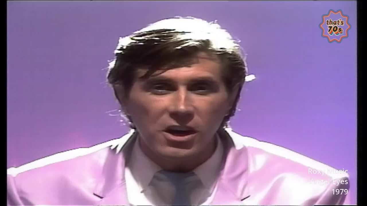 A man with feathered hair sings into the microphone. He wears a pale pink suit jacket over a white shirt and tie.