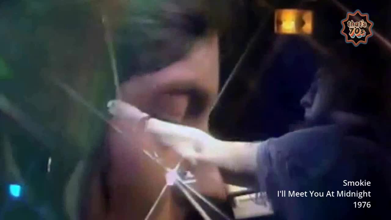 A man's hand, pale and slender, reaches out to touch another's arm. The stage lights cast a warm glow on the scene, hinting at a performance from the mid-70s.