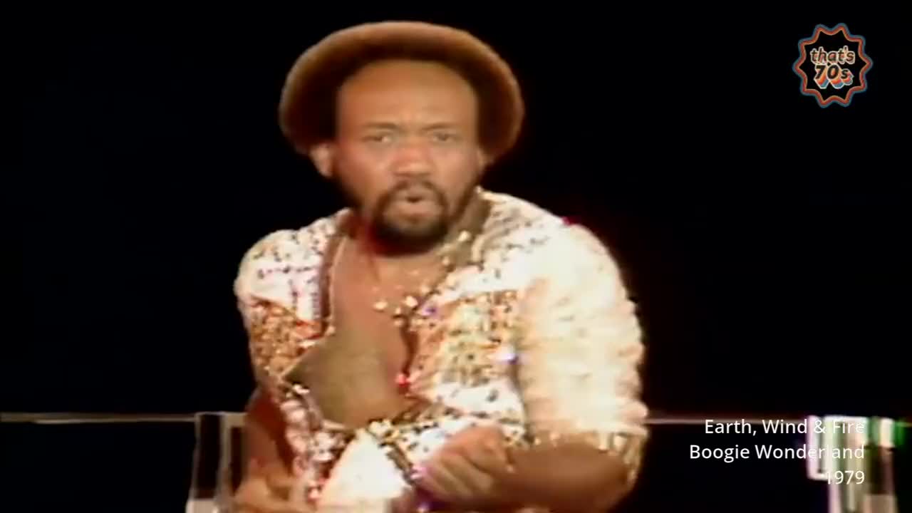 A man with a brown afro and beard, wearing a shimmering, patterned shirt, sings with his hands clasped. The stage is dark, with a hint of a railing in the foreground.