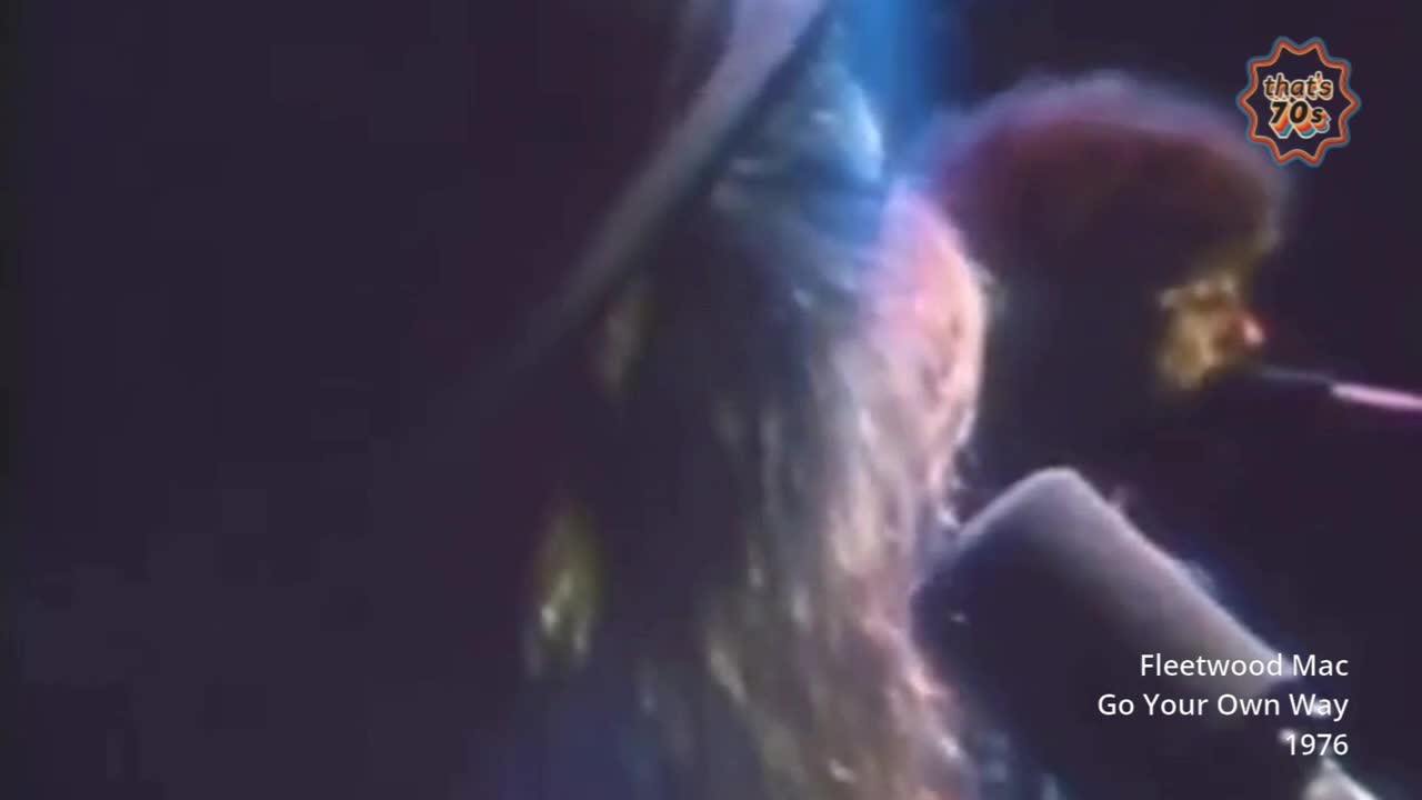 A woman with long, blonde hair, illuminated by stage lights, sings into a microphone. Beside her, a man with dark, curly hair plays an instrument, his face partially obscured by shadow.