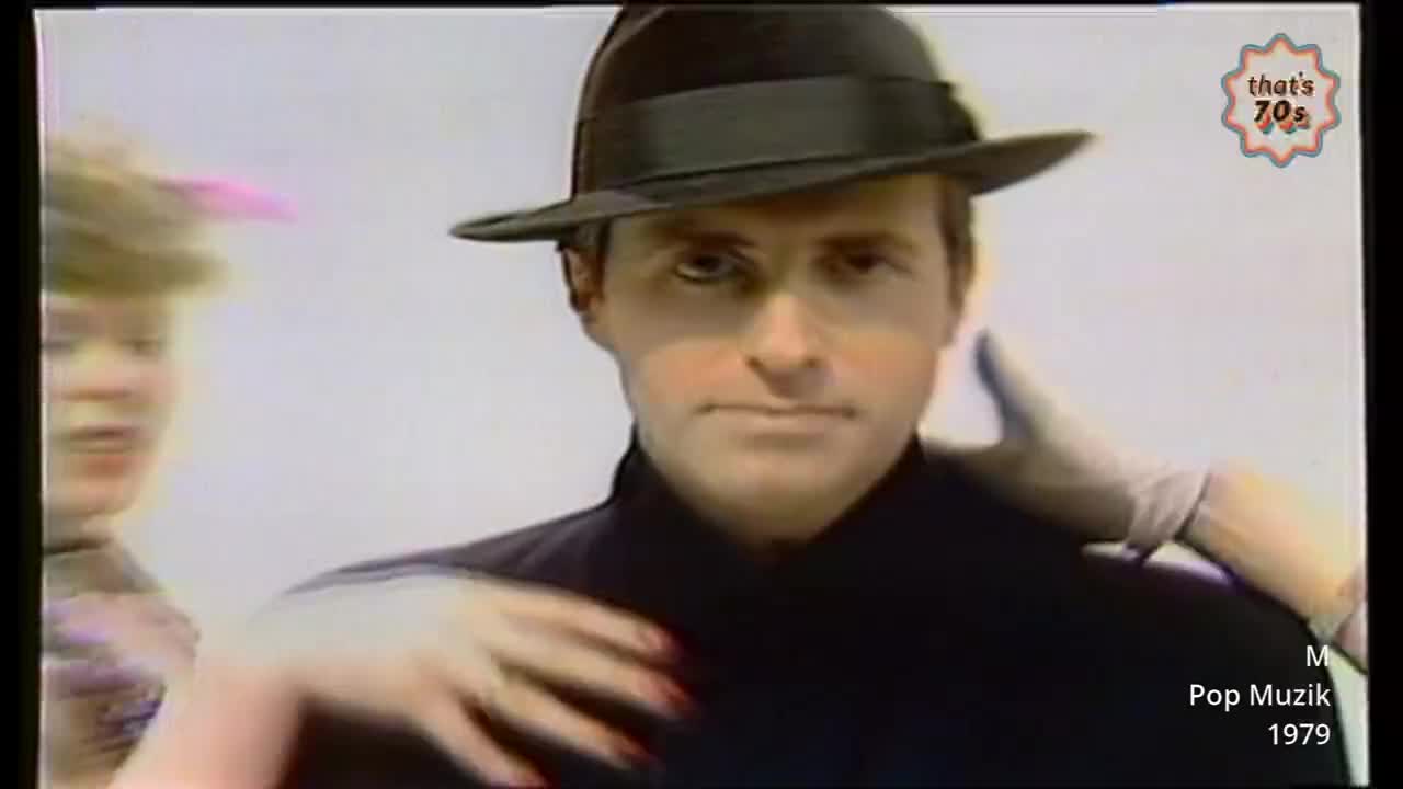 A woman with blonde hair and a pink feather in her hat leans in from the left, her hand reaching towards a man in a fedora. His eyes are fixed forward as a gloved hand rests on his shoulder.