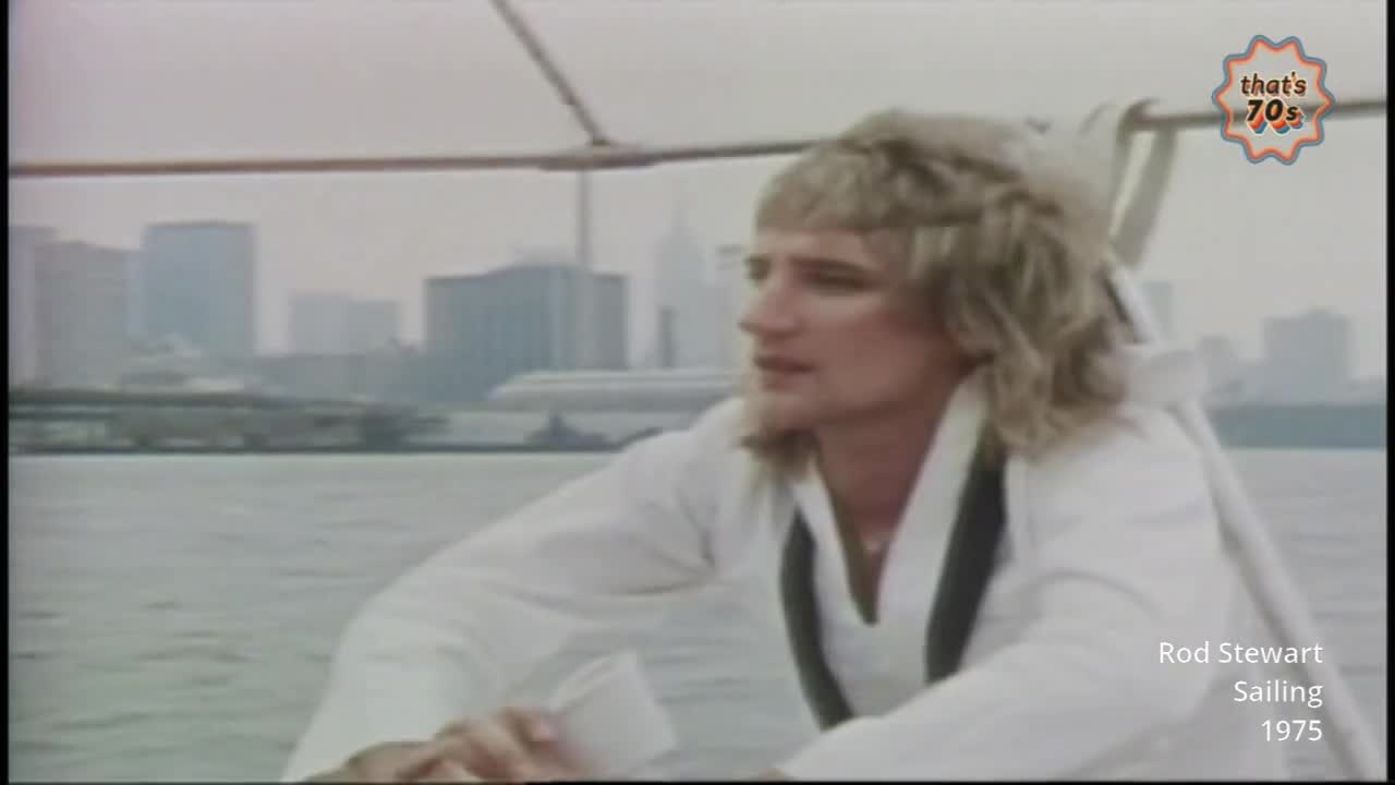 A man with shaggy blonde hair, wearing a white shirt and dark vest, looks out over the water. A hazy cityscape with tall buildings and an airplane sits in the background.