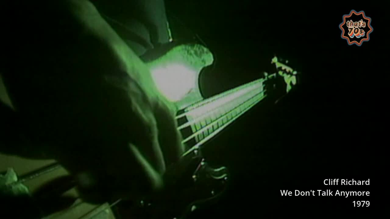 A hand moves across the fretboard of a bass guitar, illuminated by a stark green light. The strings shimmer under the glare as the musician plays.