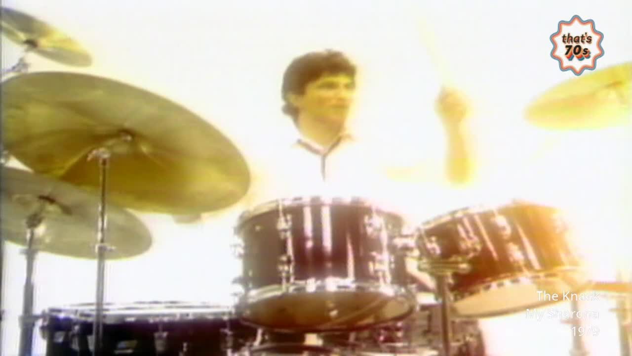 A young man in a white polo shirt is hammering away at a drum kit, his sticks a blur against the cymbals. The whole scene is bathed in a bright, hazy light, typical of a 70s music video.