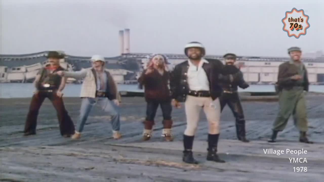 Six men in distinctive 70s outfits are performing a synchronized dance on a gritty, industrial waterfront. The man in the center, wearing a white helmet and leather jacket, leads the energetic moves.