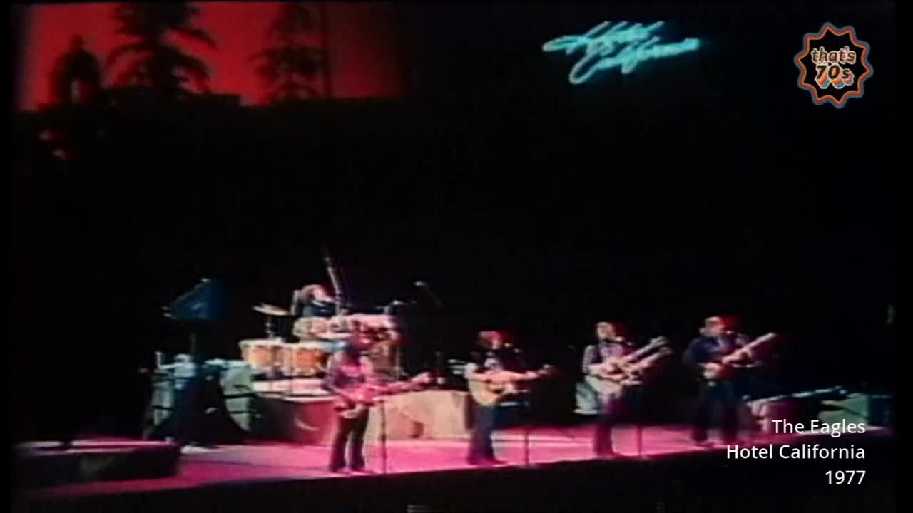 The band stands on stage, bathed in pink light, as a neon sign spelling "Hotel California" glows above them. Five men play their guitars, their silhouettes sharp against the dark backdrop.