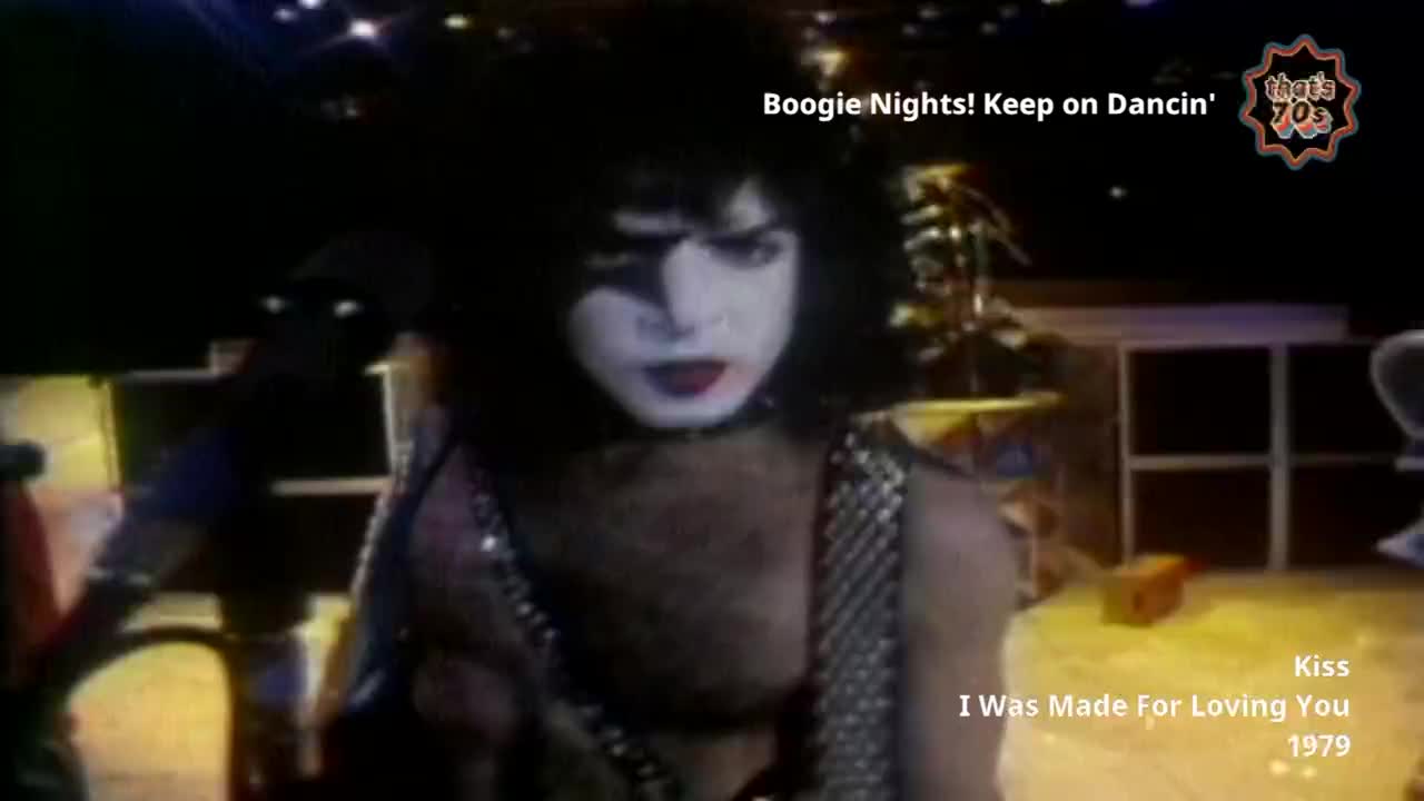 A man with white face paint and dark hair leans forward, his mouth slightly open. He wears a sparkly, sleeveless top, and the stage lights glint off the sequins.