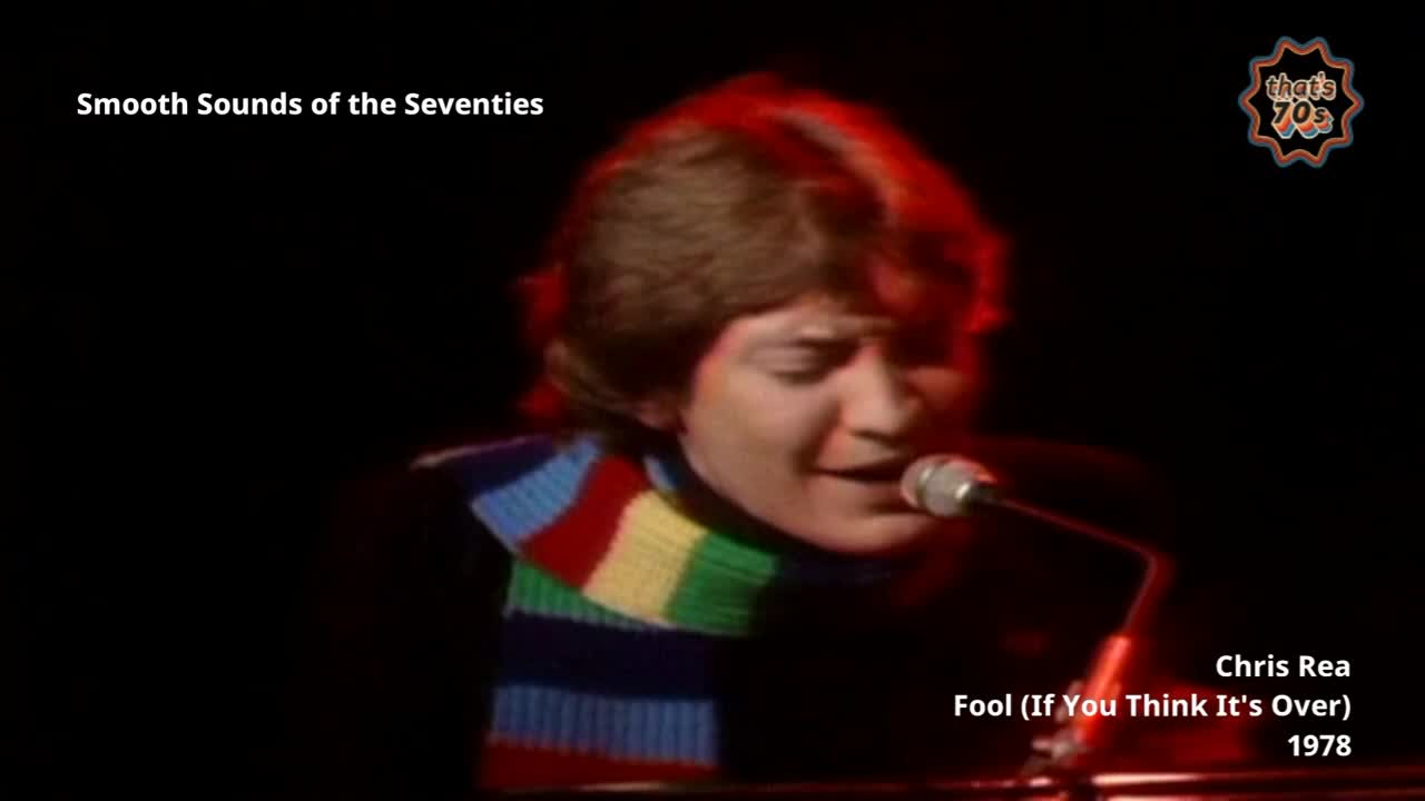 A young Chris Rea, sporting a colourful scarf, sings into a microphone. He's bathed in a warm red stage light, his face tilted upwards.