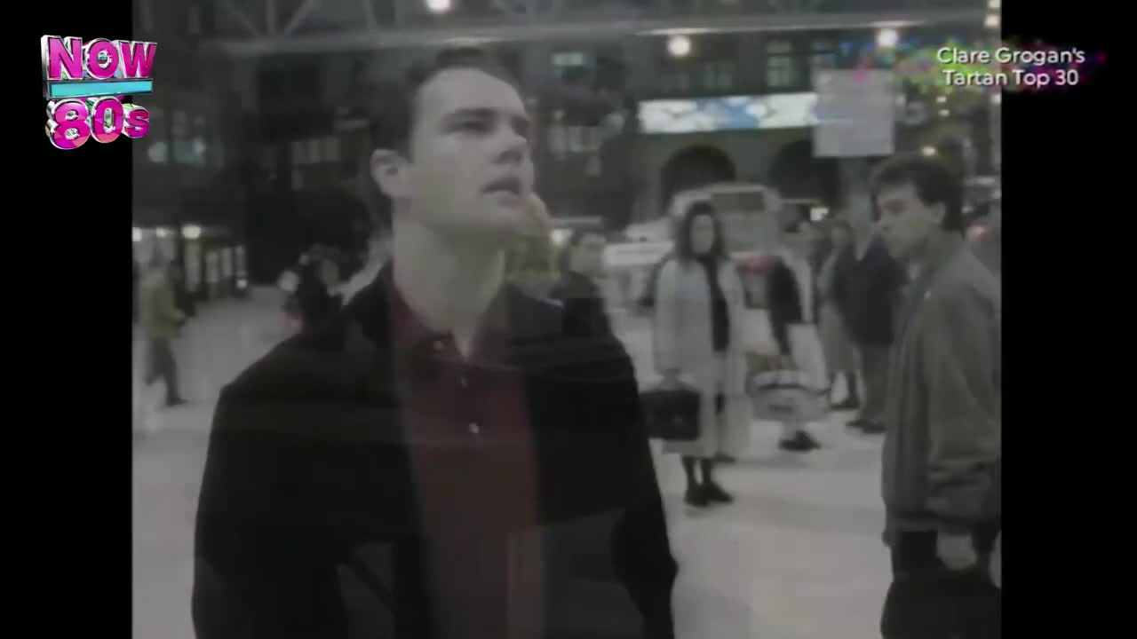 A man in a black jacket and a red shirt looks up, seemingly lost in thought, as people mill around in a bustling train station. The Now 80s logo and the "Clare Grogan's Tartan Top 30" graphic suggest this is a clip from a UK music show.
