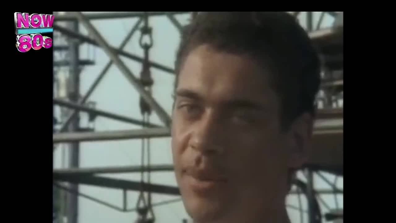 The man stares directly ahead, his expression a subtle mix of curiosity and amusement. Behind him, a metal structure rises against a pale sky, a scene from a UK music video broadcast on Now 80s.
