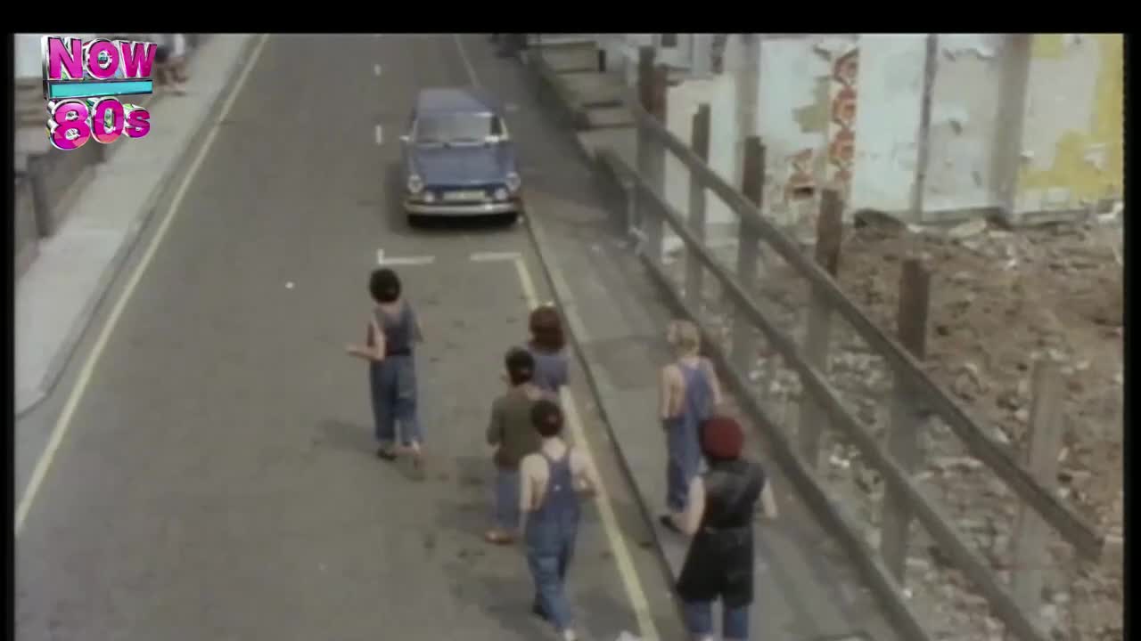 A group of children in denim overalls walk along the road, a blue car parked ahead. The scene feels like a snapshot from the UK, a moment captured from a Now 80s show.
