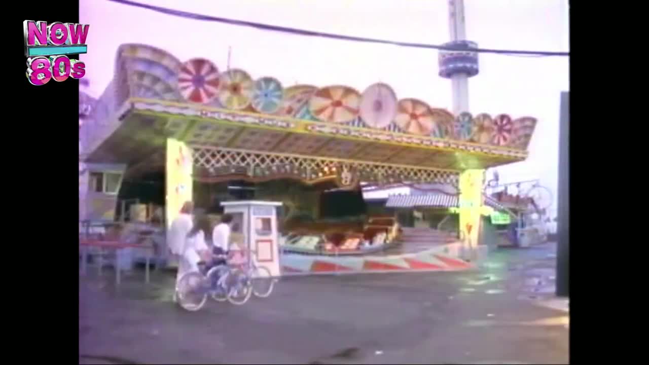 A carousel with colorful decorations spins slowly, its seats empty. A couple of people on bikes ride past the ride, near a small ticket booth.
