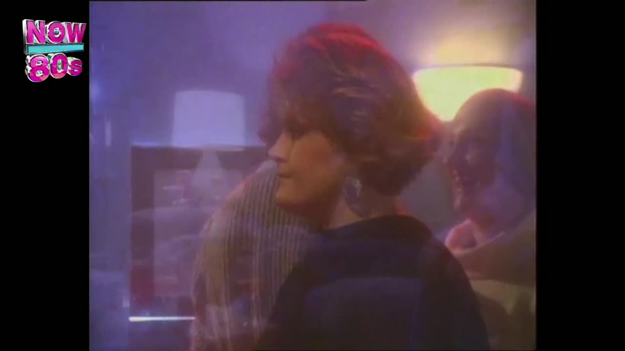 A woman with big hair turns her head, her large earrings catching the light. The scene is bathed in a purple hue, a typical look for the UK's Now 80s channel.
