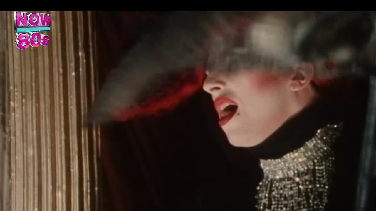 A person with heavy makeup and a beaded collar is singing, their mouth wide open. Smoke billows from their mouth, a red glow behind them.
