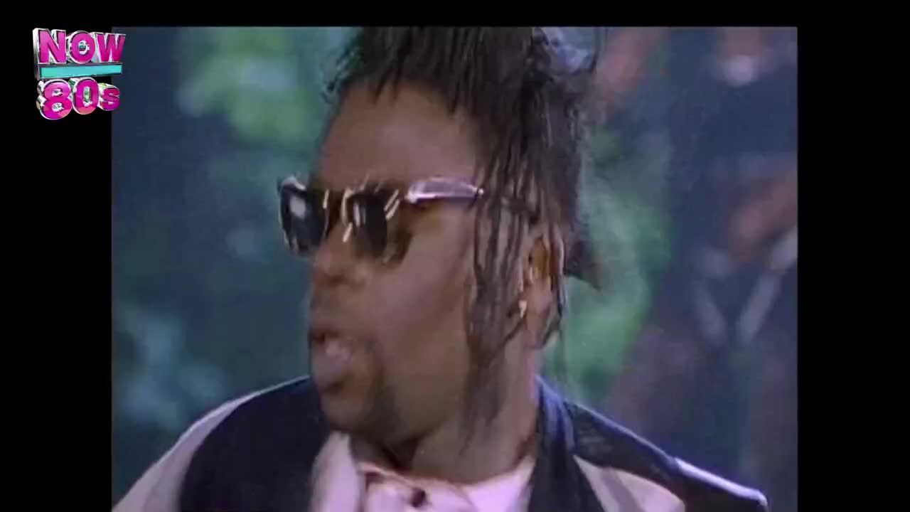 The singer, sporting sunglasses and a mohawk, is singing, his mouth open. He's wearing a pink shirt under a black and white jacket, all set against a blurred background.
