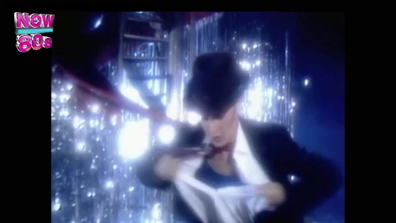 A man in a suit and hat is pulling a white cloth from his chest. Behind him, a shimmering curtain reflects the stage lights.
