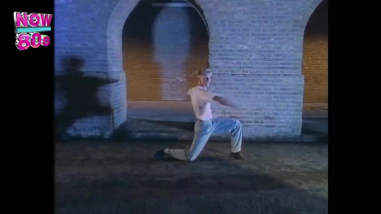 A man in a white t-shirt and jeans is kneeling, arms outstretched, in front of brick arches. His shadow stretches long across the ground, illuminated by the light behind him.
