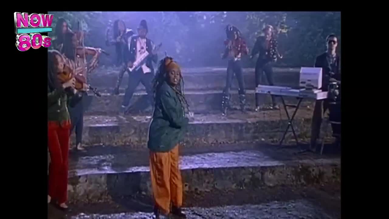 A singer in a green jacket and orange pants stands center stage, while a band plays on a set of stone steps. The musicians are dressed in various styles, from formal jackets to studded leather, as the music video unfolds.
