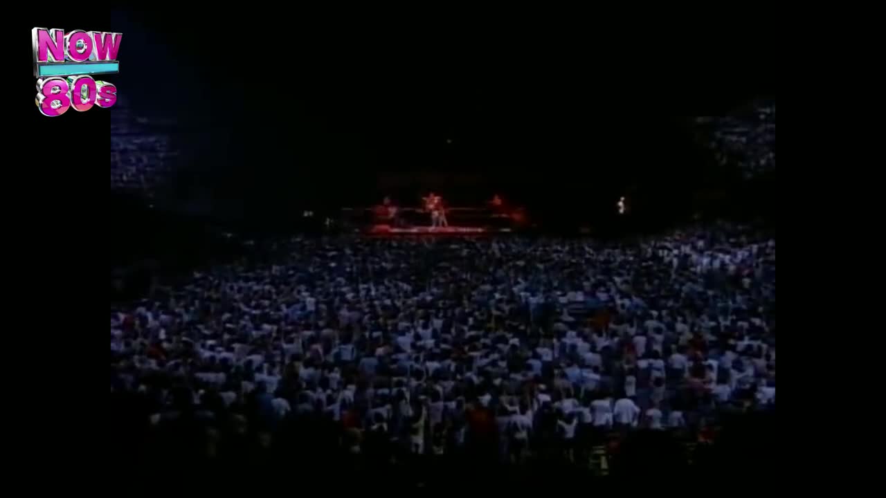 A sea of people stretches out, their faces barely visible in the dark. Up on the stage, figures are illuminated by bright spotlights.
