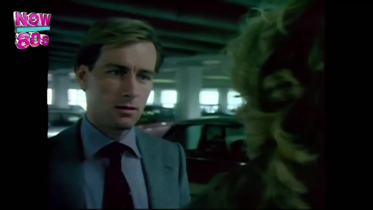 A man in a suit stares intently at someone just out of frame. The scene is set in a brightly lit parking garage, likely from a UK television show of the 1980s.
