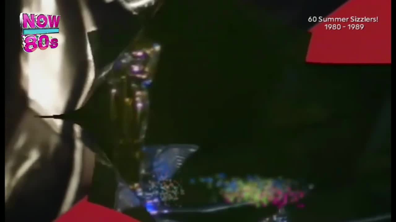 A shimmering, multifaceted shape spins against a dark backdrop, with a burst of colorful light trailing behind it. The "Now 80s" logo is visible, and the screen is advertising "60 Summer Sizzlers!" from the 1980s.
