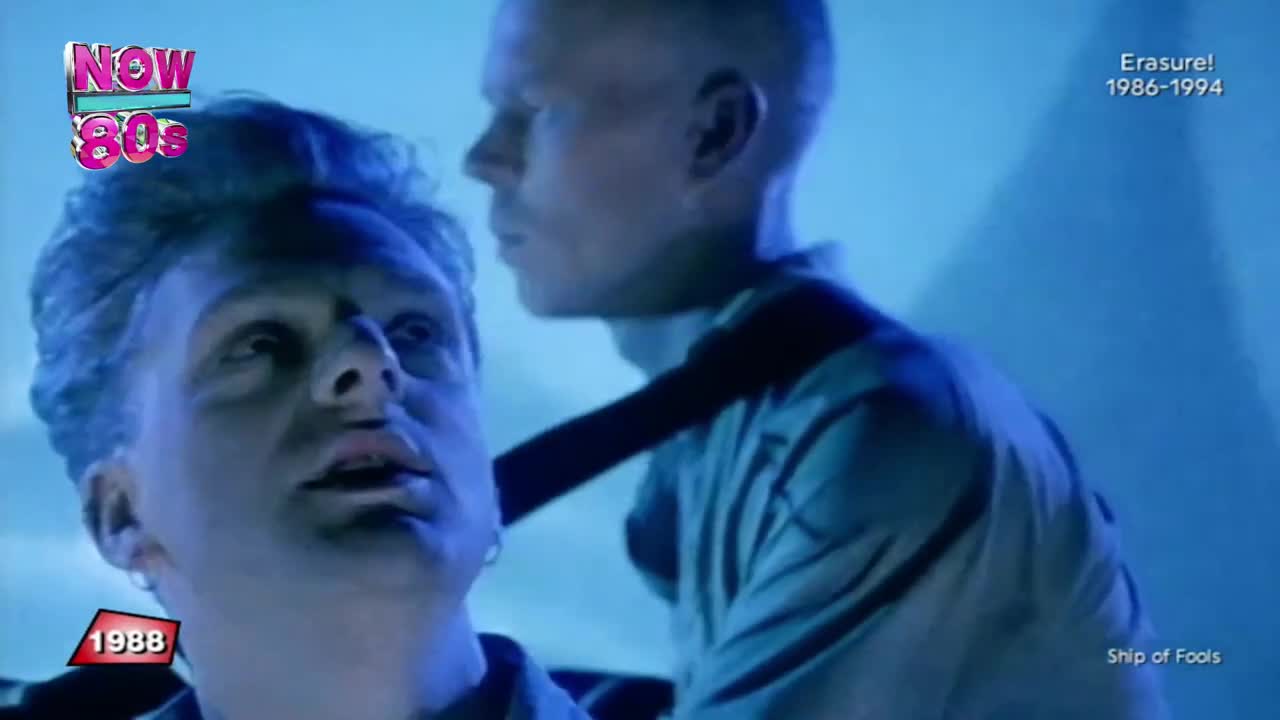 One man is leaning in, his face close to another's, a dark strap across their shoulders. The scene is bathed in blue light, with a vintage logo in the corner.
