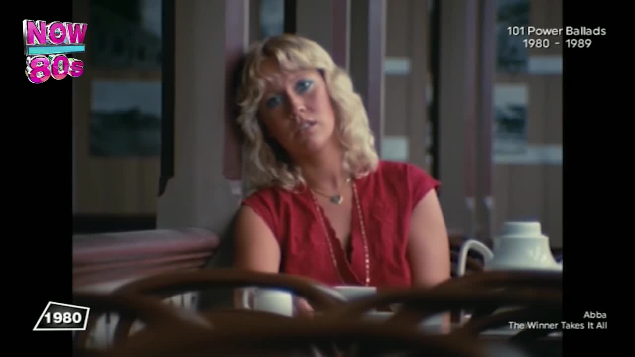 A woman with blonde hair leans against a wall, looking pensively towards the camera. She wears a red top and a long necklace, a coffee cup sits next to her.

