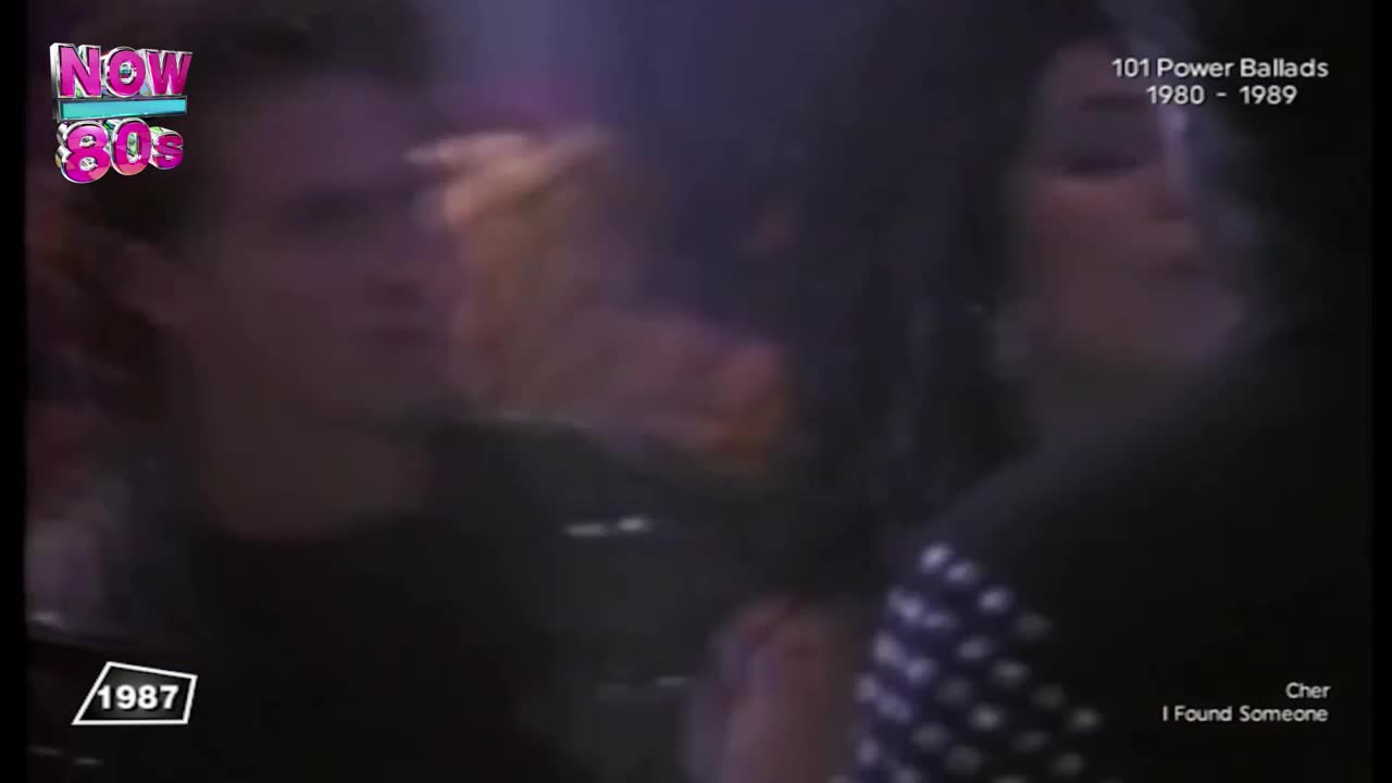 A man and a woman are in the dim light, the woman wearing a dark top with a white pattern. The screen shows the Now 80s logo, and the year 1987 is visible.

