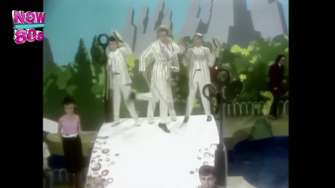 Three men in white striped suits are dancing on a raised platform, each holding a large, black ring. A woman in a pink top and black skirt stands to the side, watching.
