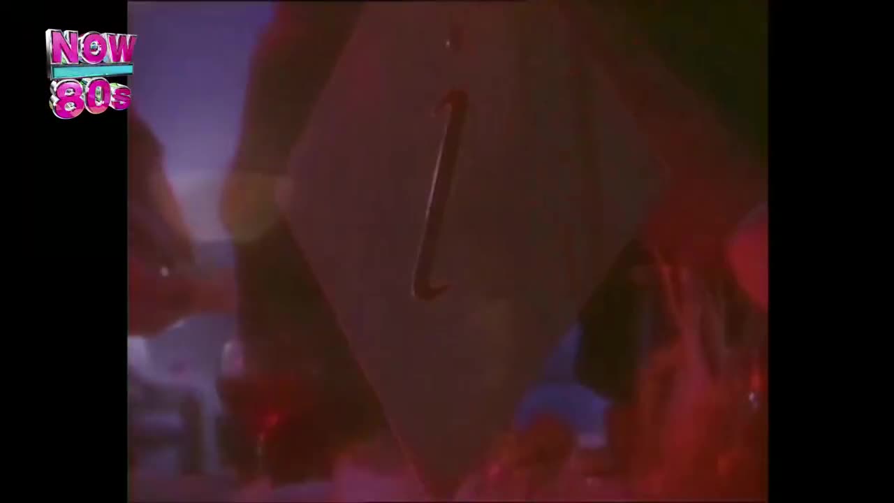 A large, white shape with a curved handle is being moved around the stage. The Now 80s logo is in the corner, and the lighting is a mix of red and blue.
