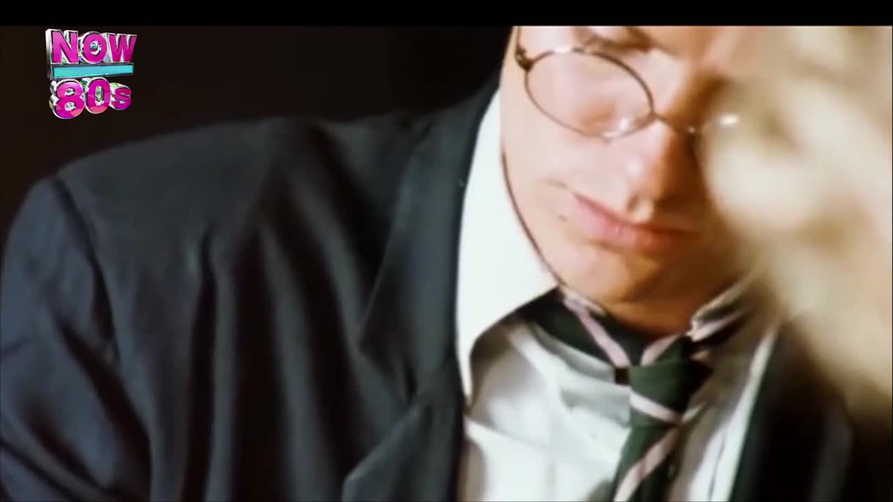 A man in a suit and glasses stares down, his tie askew. The logo for Now 80s sits in the corner, a reminder of the era.
