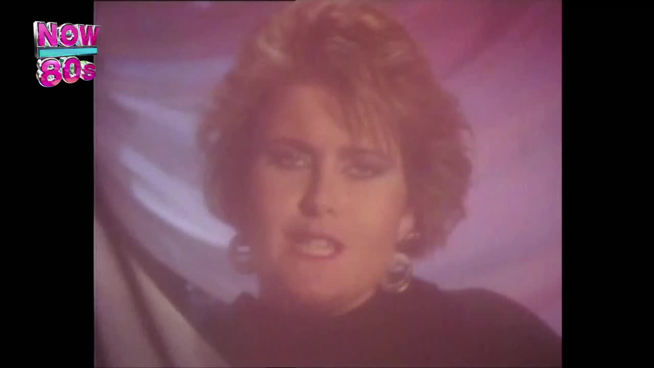 A woman with big hair and hoop earrings is singing, her face lit by a soft pink glow. The "Now 80s" logo sits in the corner, a familiar sight on UK television.

