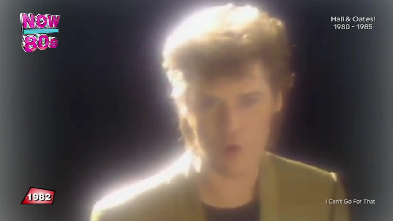 A man in a light green jacket is singing, his face illuminated by a bright glow. The "Now 80s" logo and a year are in the corner, suggesting a music video from the United Kingdom.
