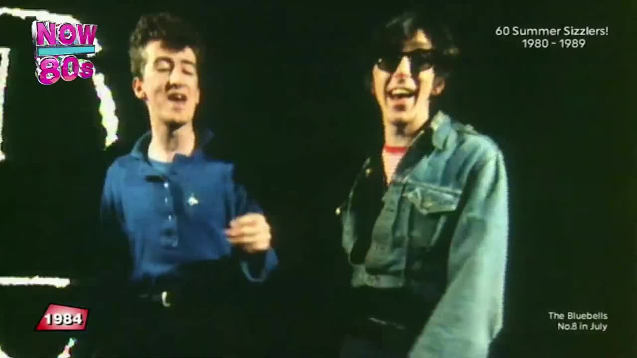 Two men are singing, both grinning at the camera as they move. One is in a blue polo shirt, the other in a denim jacket and sunglasses.
