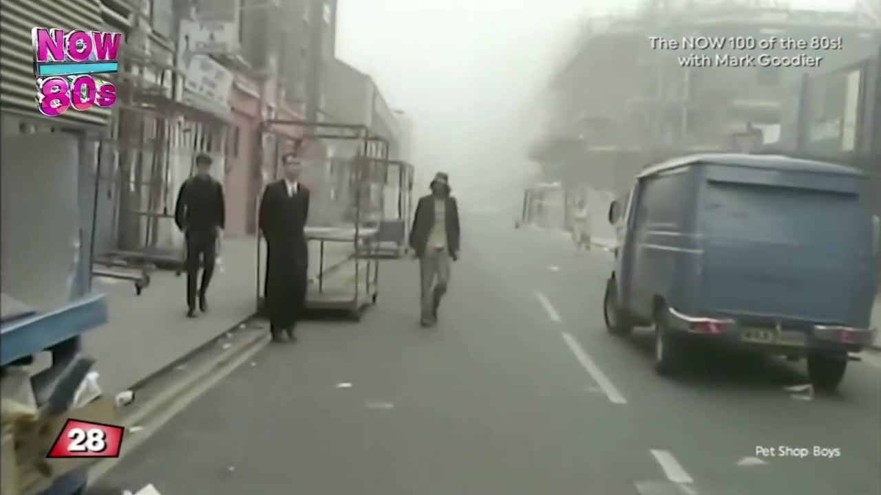 Three men walk down a foggy street, one in a long coat, another in a jacket and hat. A blue van moves slowly past them.
