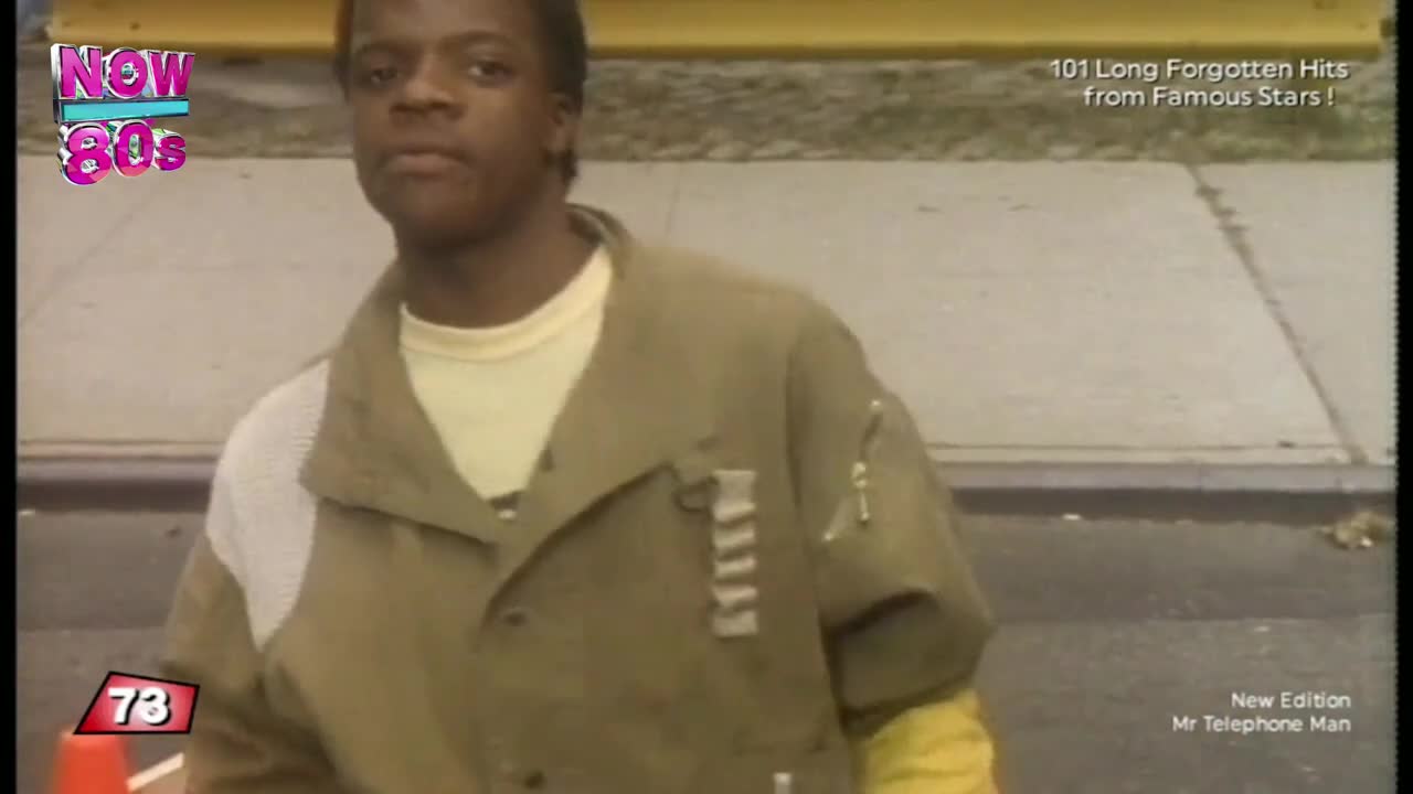 A young man in a khaki jacket stands on what looks like a street corner, staring directly at the camera. The "Now 80s" logo and a song title card suggest a music video from the United Kingdom.
