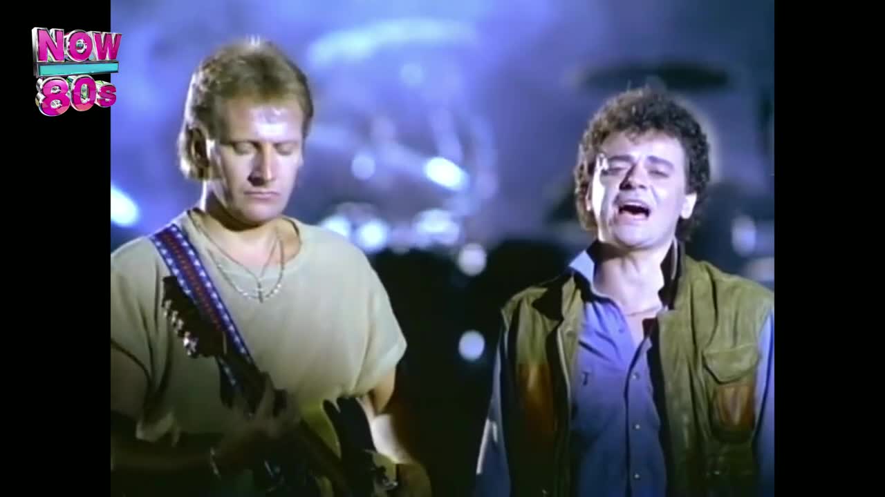 The guitarist, in a pale green shirt, strums his instrument, while the singer beside him belts out the song with his mouth wide open. A blue-tinged background provides the backdrop for this performance.
