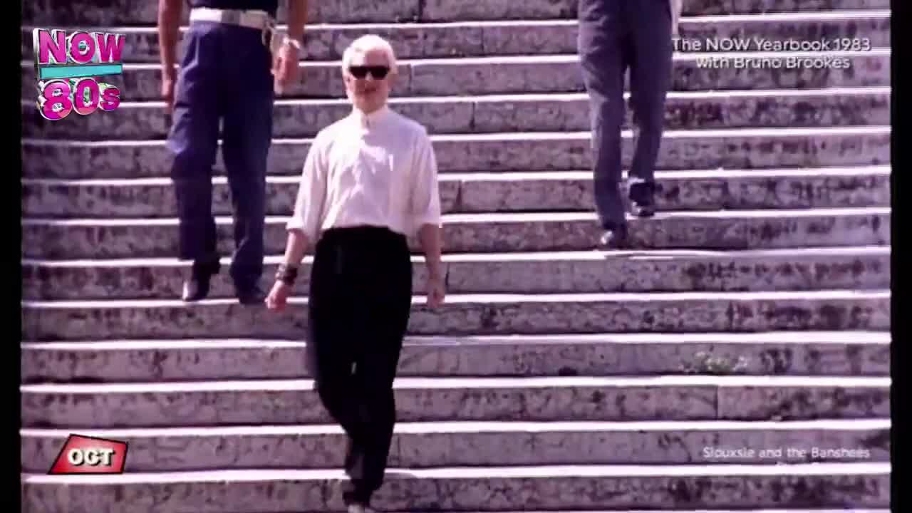 A man in sunglasses and black pants descends the steps, followed by two others. It's a scene from a NOW 80s show in the United Kingdom, with the band Siouxie and the Banshees featured.
