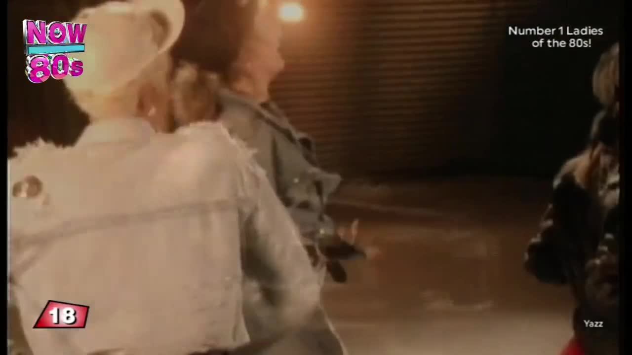 A woman in a white denim jacket is turning, her back to the camera, while another woman in a denim jacket is dancing. The scene is lit dimly, a Now 80s logo in the corner.
