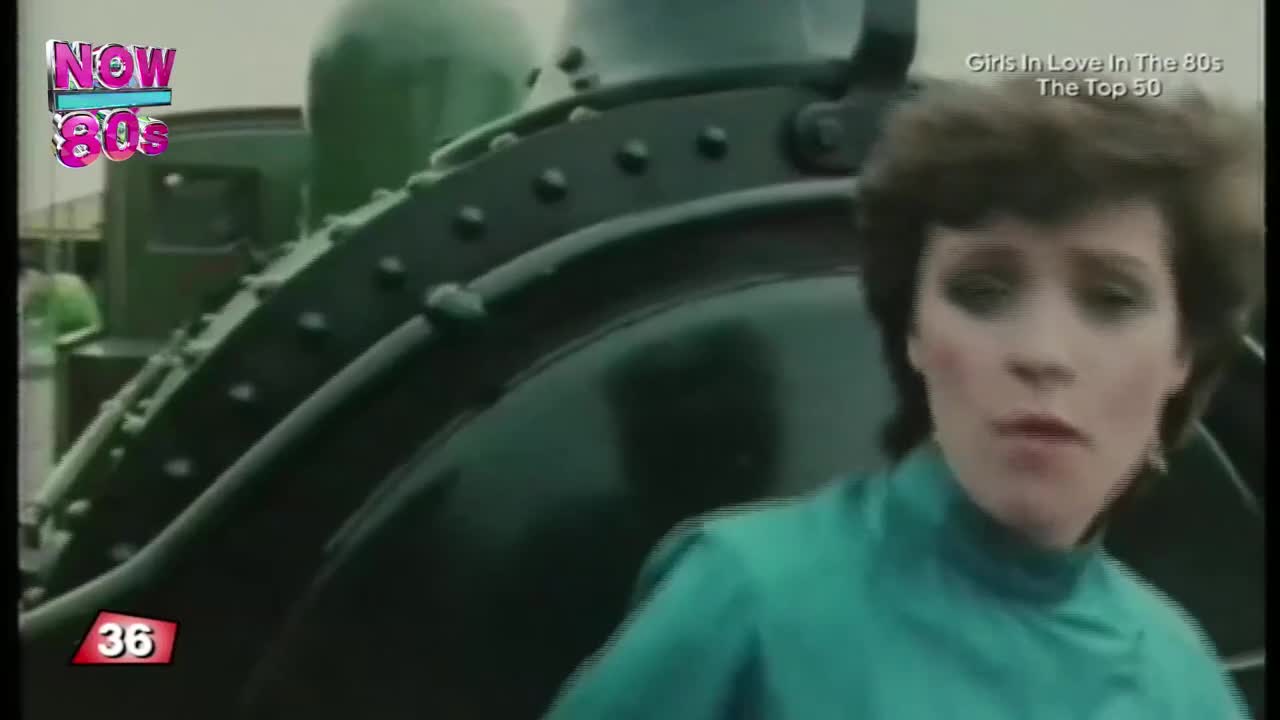 A woman in a teal top stands in front of a dark green train, singing. The Now 80s logo is visible, and it's a music video from the United Kingdom.
