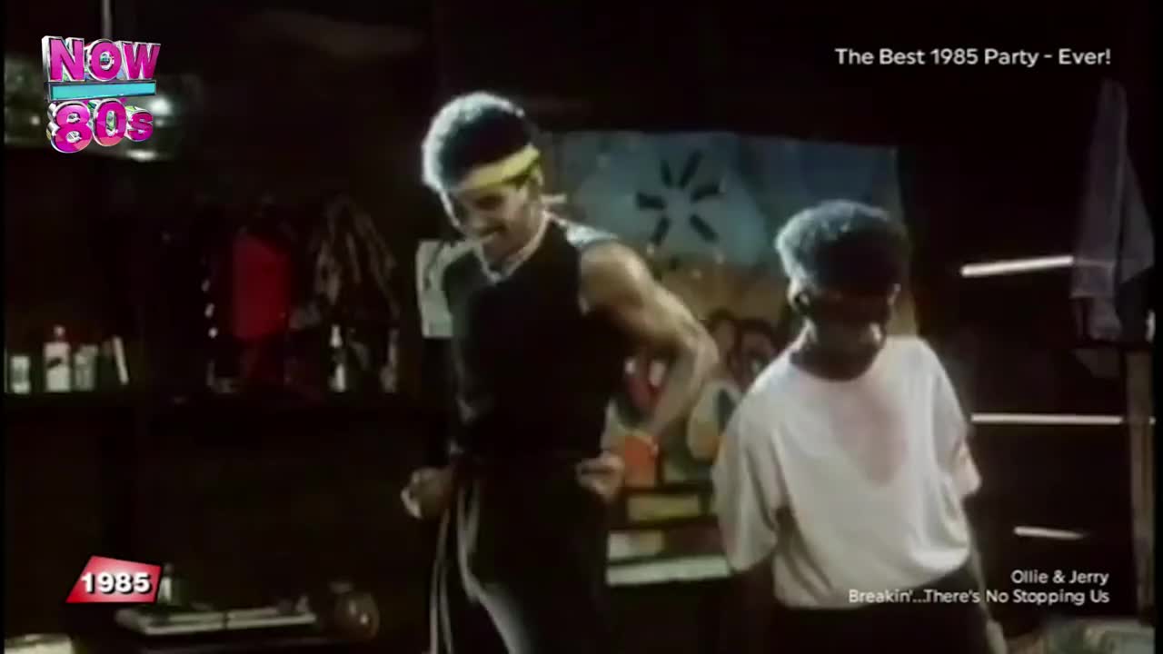 Two men, one in a black vest and a yellow headband, are dancing in a dimly lit room. The other man, wearing a white t-shirt, bobs his head to the music as they move to the beat.

