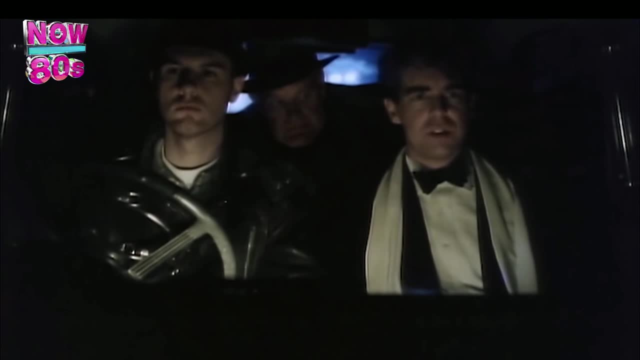 Inside a car, three men are illuminated by the interior lights, one in a leather jacket at the wheel, another in a tuxedo, and a shadowy figure in between. The scene feels like a clip from a music video, probably from the UK, playing on Now 80s.
