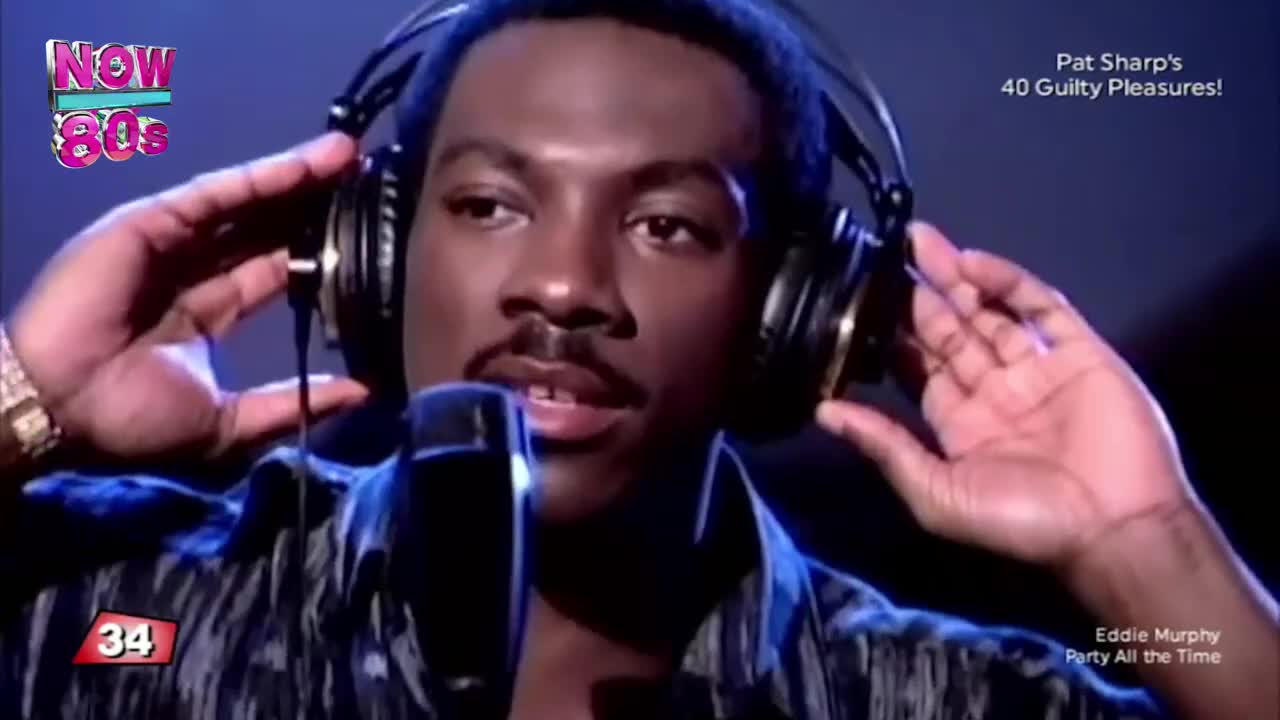 Eddie Murphy, in a patterned shirt, holds his headphones, the microphone close to his mouth. The "Now 80s" logo is visible, and the screen displays "Party All the Time."

