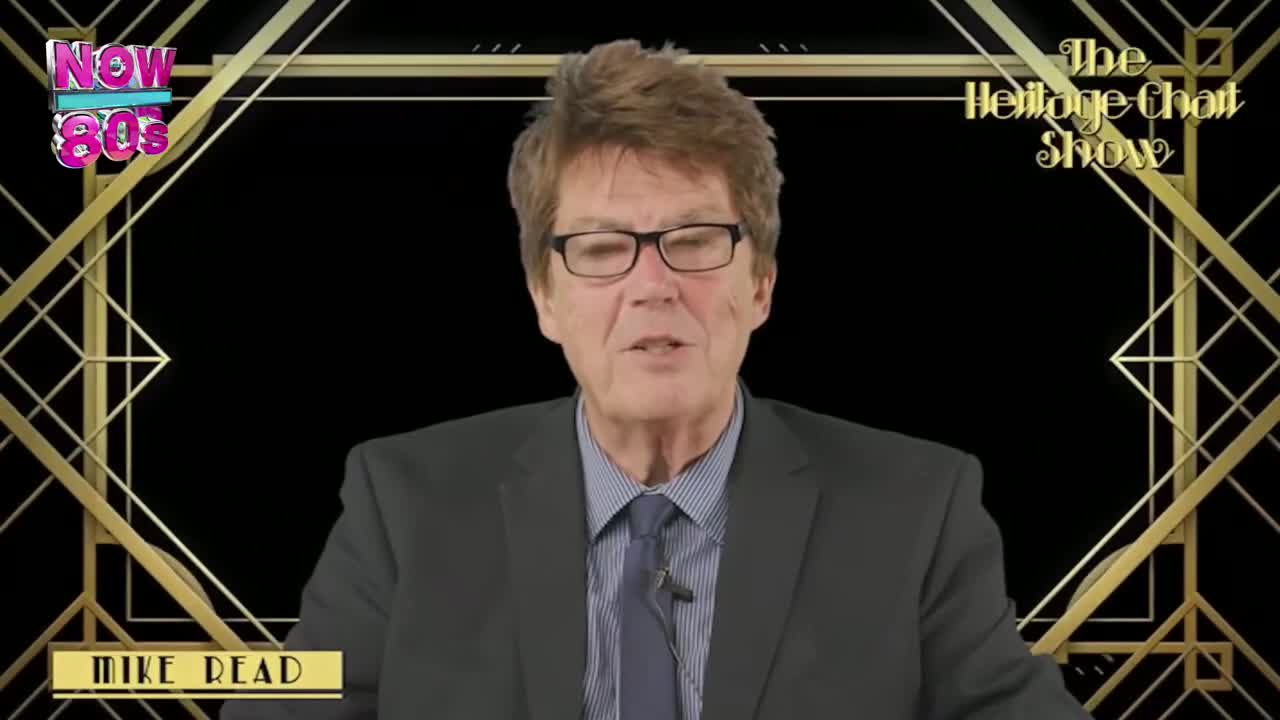 A man in a suit and tie, wearing glasses, is speaking directly to the camera, a small microphone clipped to his lapel. The background is a black screen with a gold art deco design, and the "Now 80s" logo is visible.
