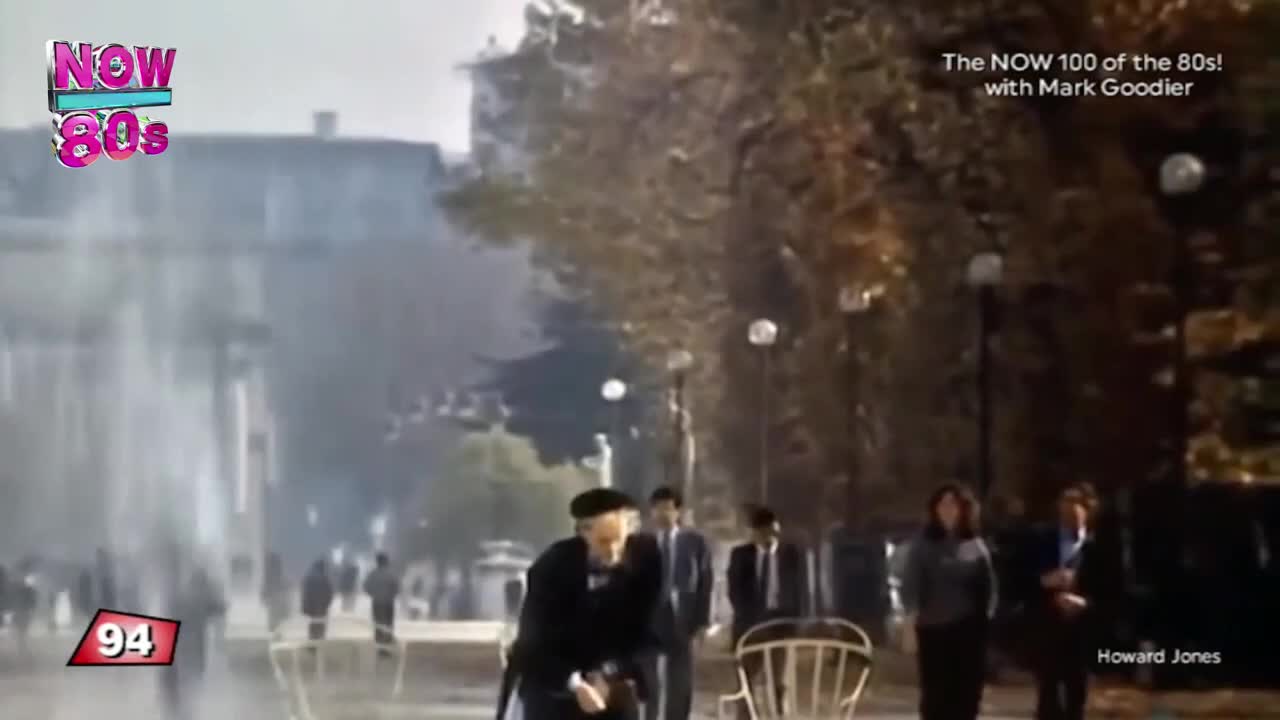 A man in a dark coat and a beret walks towards the camera, followed by a group of people in suits. Behind them, a water feature sprays into the air, creating a misty effect.
